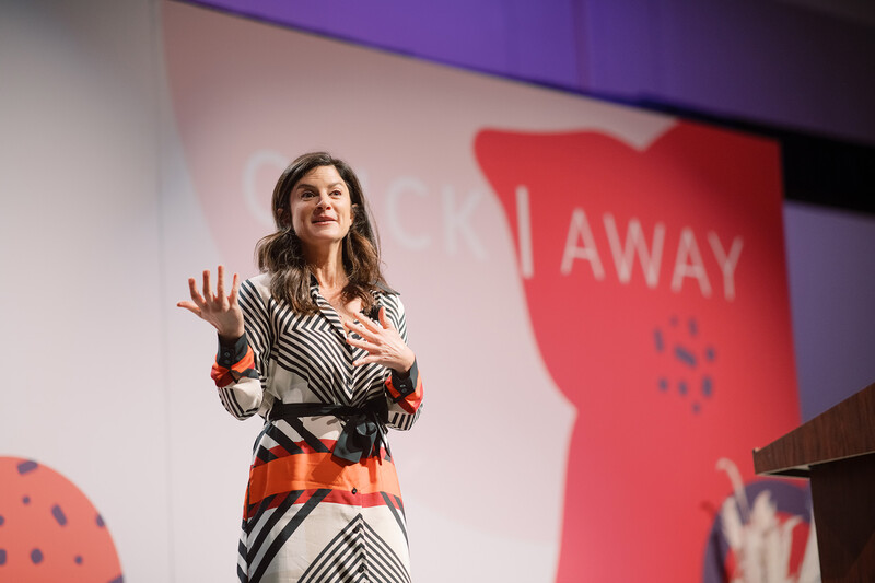 Woman speaking on stage with animated hand gestures, wearing striped and coral patterned clothing.