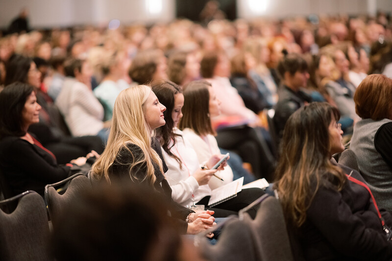 Audience members seated in an auditorium, with a woman in foreground holding a notebook and pen.