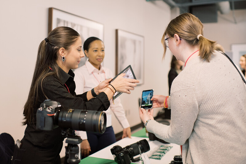 Woman with professional camera demonstrates smartphone photo to female client at product display.
