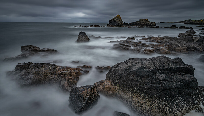 Rocky coastline with smooth water and dramatic cloudy sky at dusk or dawn.