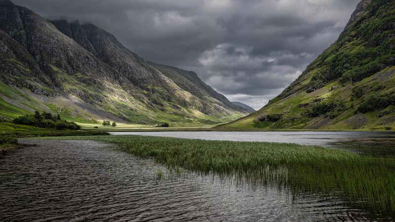 Mountain valley with a calm lake and reeds in the foreground, steep green slopes on either side, and dramatic overcast sky.