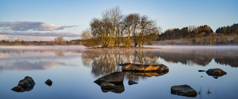 Misty lake at dawn with dark rocks in foreground and tree-lined island reflecting in still water.