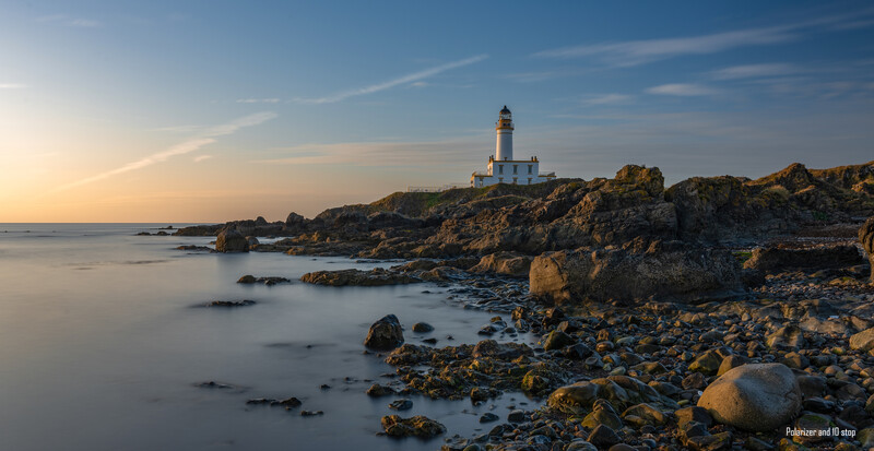 Lighthouse on rocky headland at golden hour with smooth water and scattered boulders in foreground.
