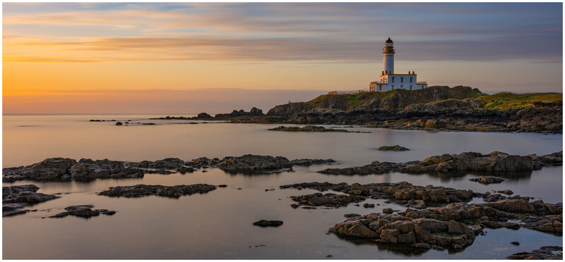 Lighthouse on a rocky headland at sunset with calm sea and dark rocks in foreground.
