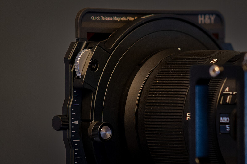 Close-up of a large format camera lens with quick-release magnetic filter system mounted on a dark background.