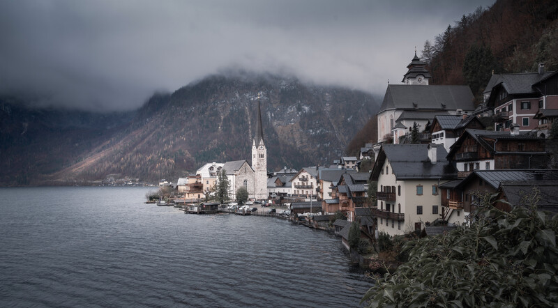 Alpine lakeside village with church spire nestled between forested mountains shrouded in mist.