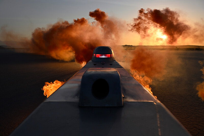 Drag racing vehicles launching with intense flames and orange dust clouds trailing behind on a desert track at sunset.
