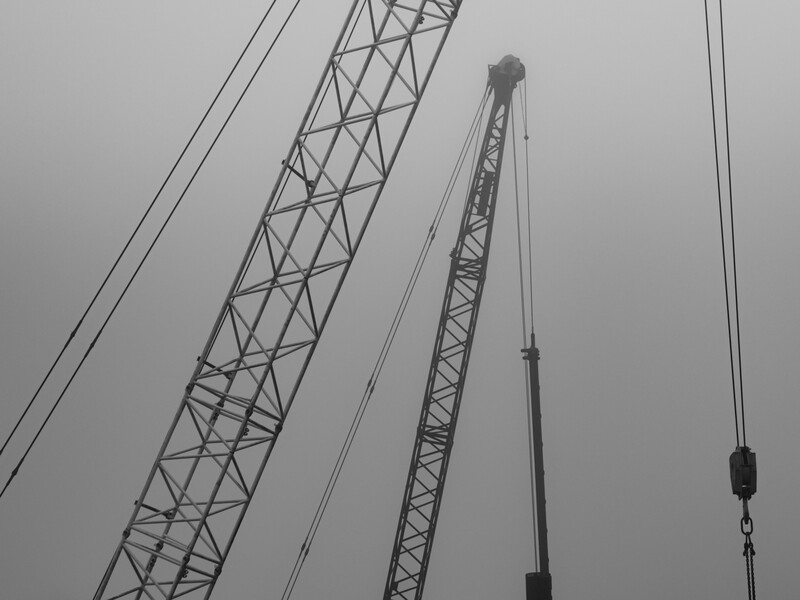 Industrial crane structure with triangular lattice boom and cable rigging against overcast sky.