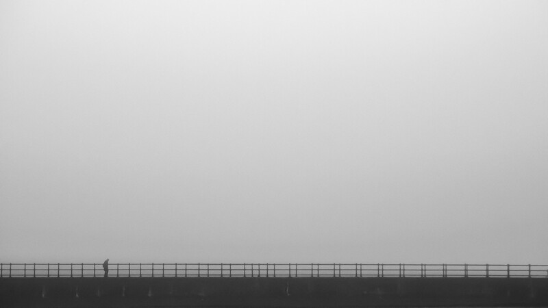 Solitary figure standing on a long horizontal railing or fence against an overcast sky.