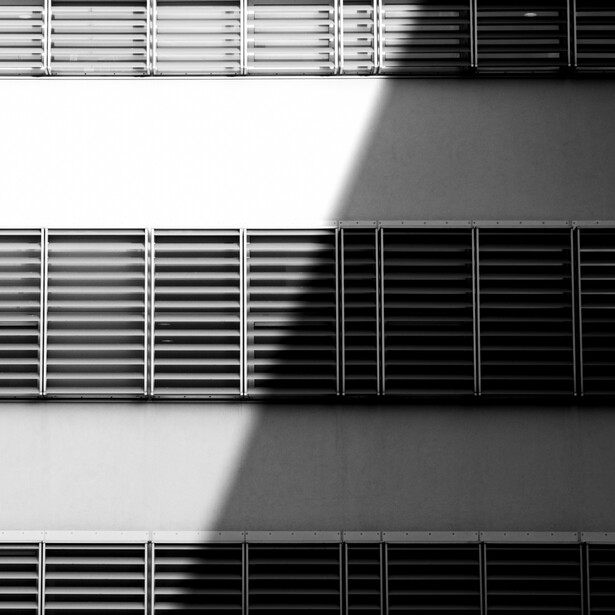 Minimalist black and white architectural photograph showing three rows of horizontal louver windows on a building facade with dramatic shadow and light division.