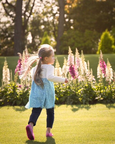 Young child running on a lawn toward blooming flower beds in golden afternoon light.