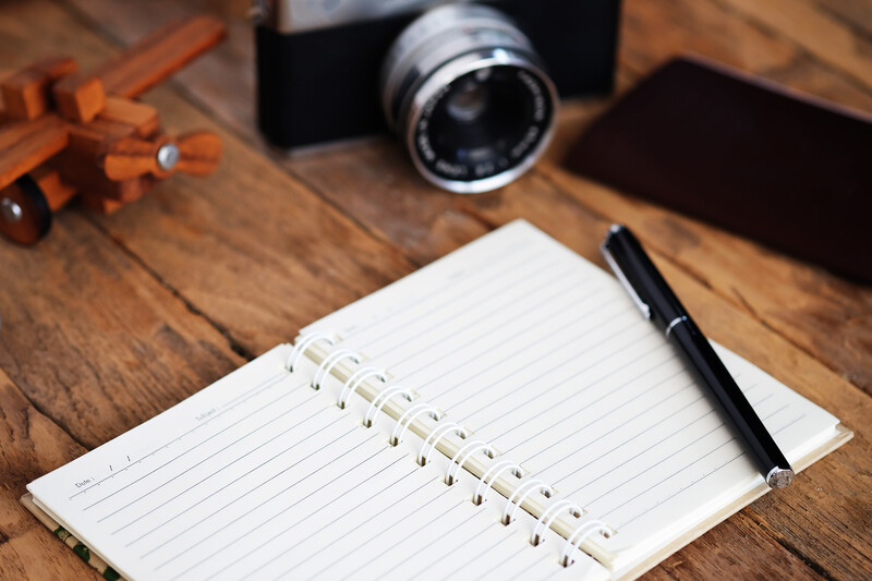 Overhead flat lay of a spiral-bound notebook with lined pages, black pen, vintage camera lens, and wooden camera body on warm wood surface.