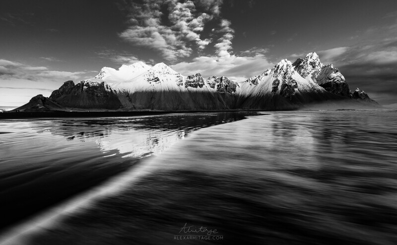 Snow-capped mountain range reflected in still water under dramatic sky.