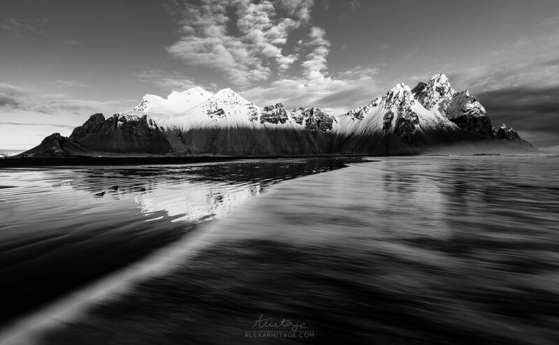 Snow-capped mountain range reflected in still water under dramatic sky.