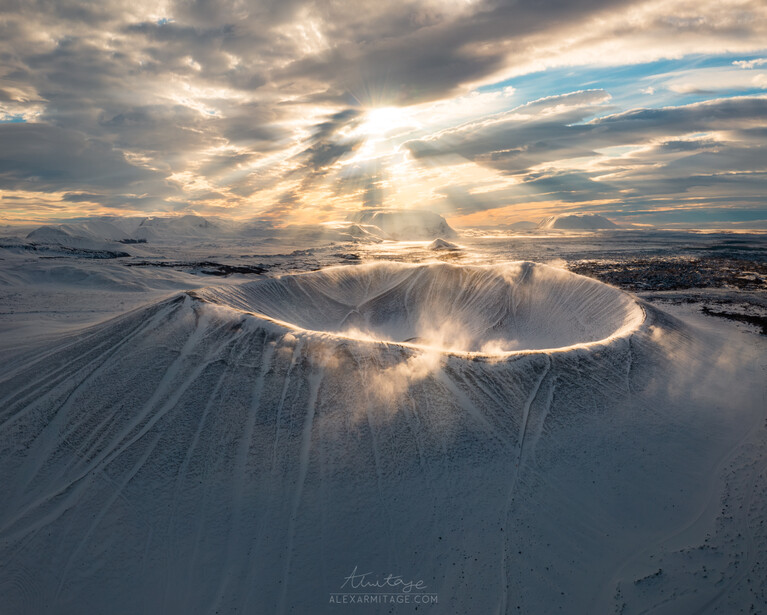 Snow-covered volcanic cone with dramatic crepuscular rays breaking through clouds at sunset.