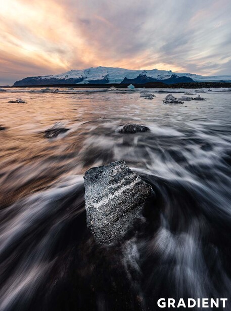 Boulder in turbulent seawater during golden hour with mountains in distant background.
