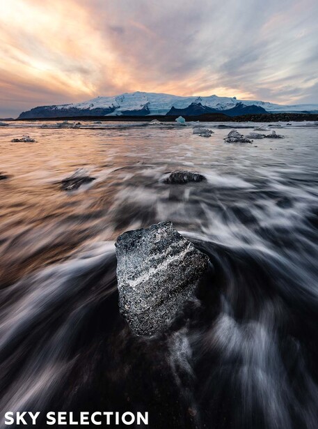 Boulder in turbulent water with mountains and dramatic sunset sky in the background.