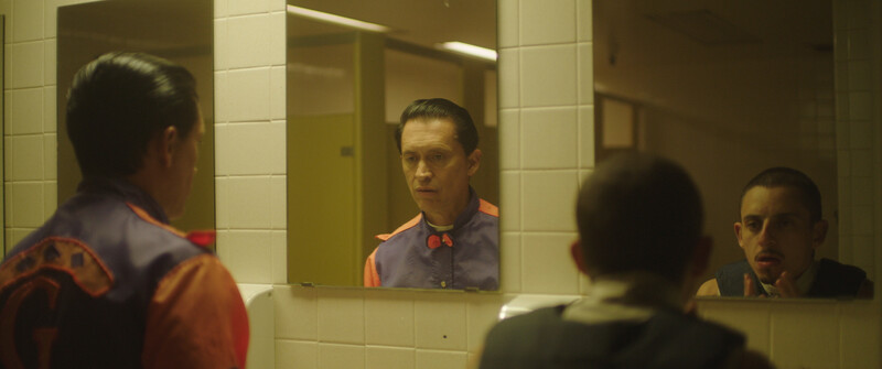 Man in orange-trimmed shirt reflected in bathroom mirror, with another person visible in foreground.