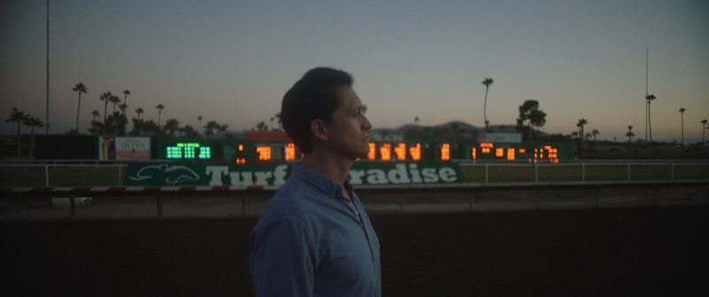 Man in blue shirt standing in profile at dusk with illuminated scoreboard and palm trees behind him.