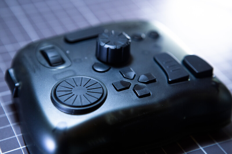 Close-up of a black gaming controller with analog stick, buttons, and textured grip on a gridded surface.