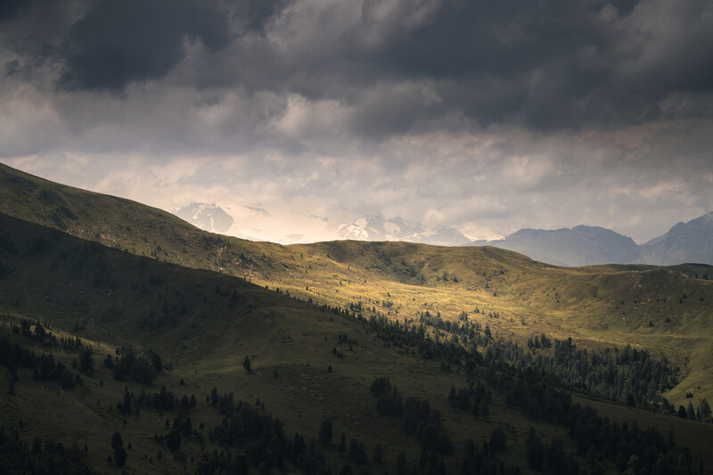 Rolling forested mountain valleys under dramatic cloud-filled sky with distant snow-capped peaks.