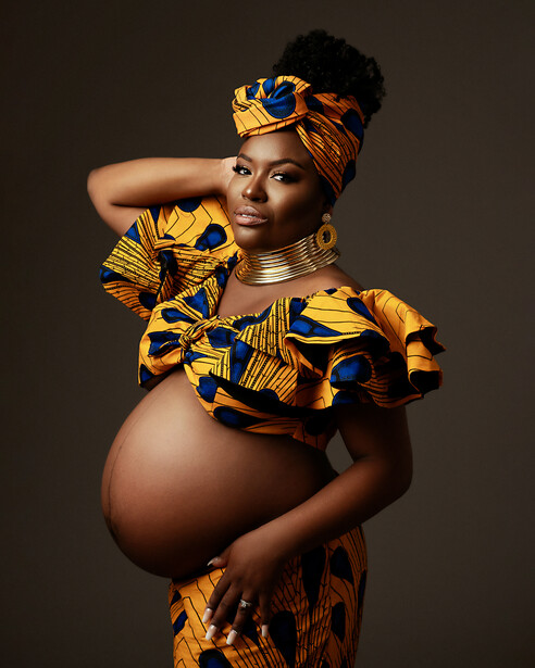 Pregnant woman in gold and black patterned outfit poses against neutral background.