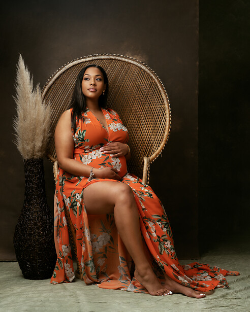 Woman in orange patterned dress seated in woven peacock chair against dark background.
