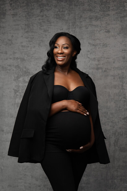 Pregnant woman in black maternity outfit against textured gray background.