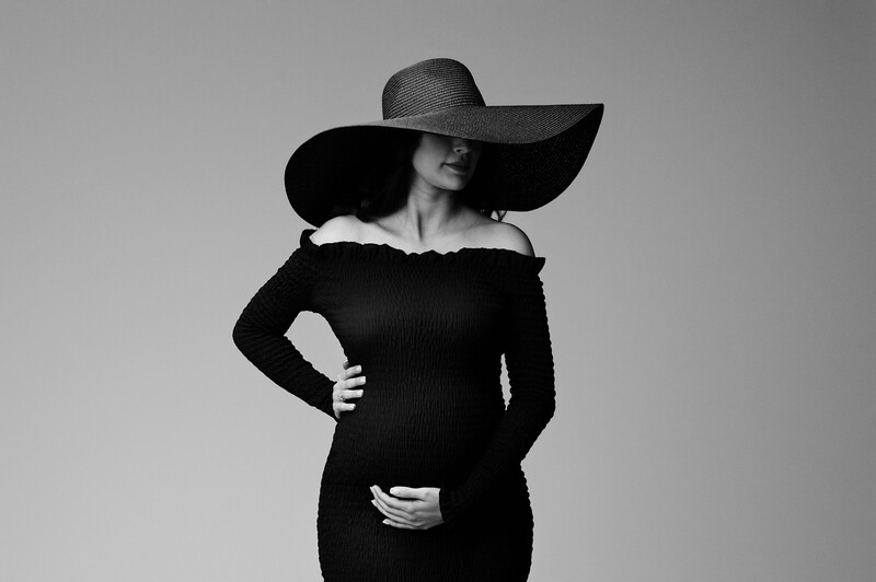Woman in black off-shoulder dress wearing a large dark wide-brimmed hat, posed against neutral background.