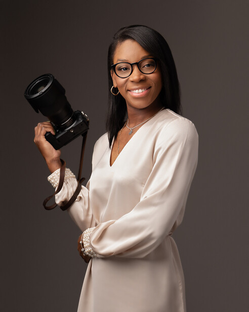 Professional photographer holding a telephoto lens while smiling at the camera against a neutral gray background.