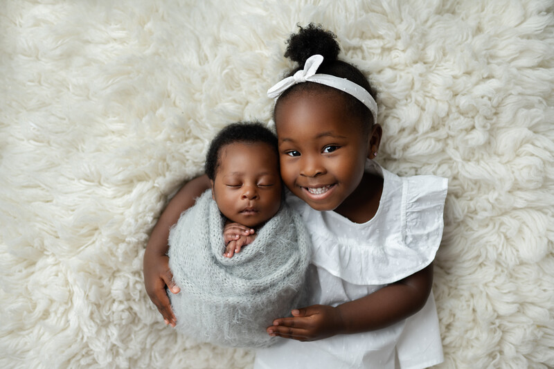 Two young children posing together on a cream-colored fuzzy background. Older child smiles at camera while embracing younger swaddled infant.