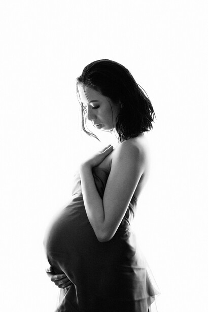 Pregnant woman in profile wearing draped fabric, posed against white background in black and white.