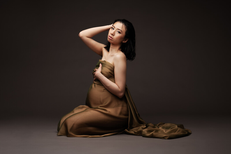 Woman in bronze silk dress seated on gray floor against dark background, hand raised to head.