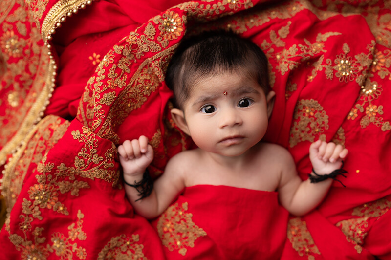 Infant lying on red fabric with gold embroidered patterns, looking directly at camera with neutral expression.