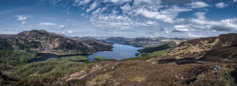 Expansive mountain landscape with a large blue lake nestled among forested valleys and rolling peaks beneath a partly cloudy sky.