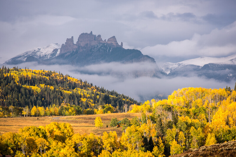 Mountain peak shrouded in mist rises above golden aspen trees and evergreen forest in autumn valley.