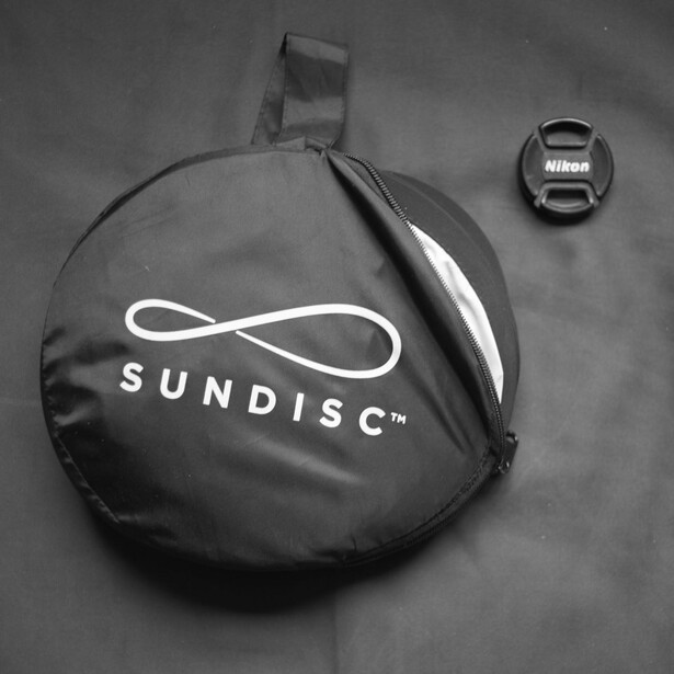 SunDisc camera lens pouch next to Nikon lens cap on gray surface.