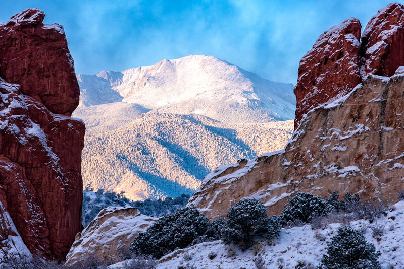 Snow-dusted red rock formations frame a distant snow-capped mountain peak under clear blue sky.