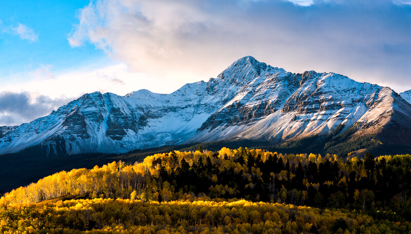 Snow-capped mountain peak above golden larch forest in autumn light.