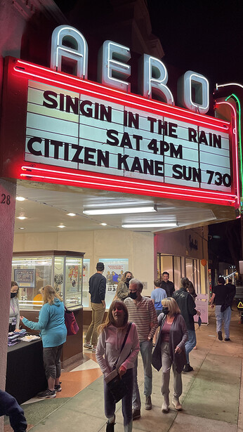 Neon-lit movie theater marquee displaying film titles and showtimes at night, with patrons visible in the lobby below.