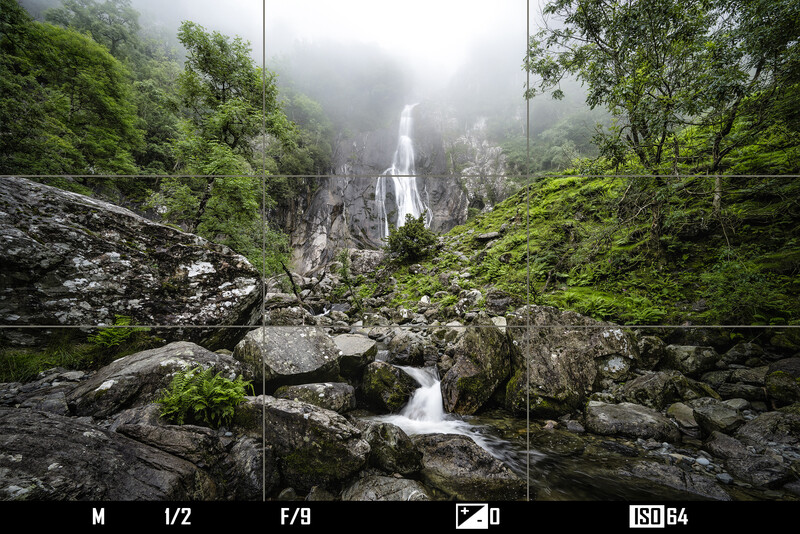 Mountain waterfall cascading through forested canyon with misty peaks in background.