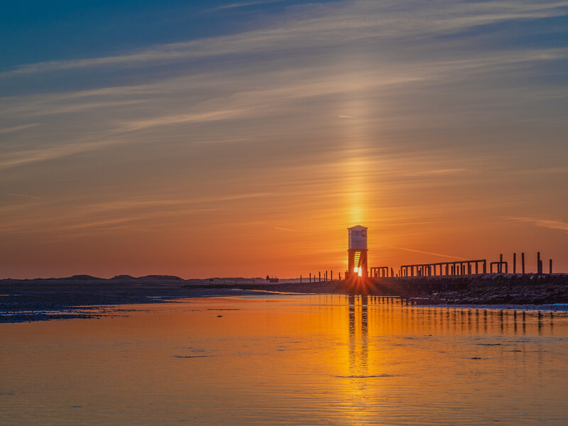 Industrial waterfront at sunset with a tall tower reflected in calm water.