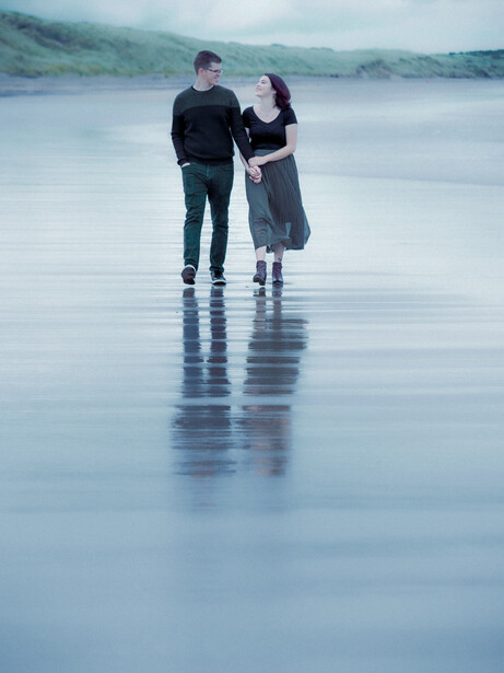 Couple standing together on a reflective wet beach or tidal flat, with misty hills in the background.
