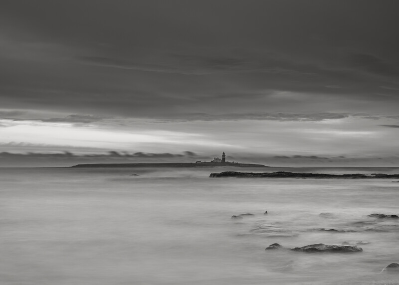 Long exposure seascape with rocks in foreground and distant lighthouse on horizon under dramatic cloudy sky.