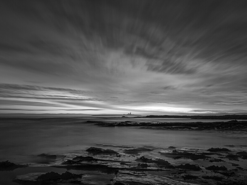 Rocky coastal rocks in foreground with calm water and dramatic sky at dusk or dawn.