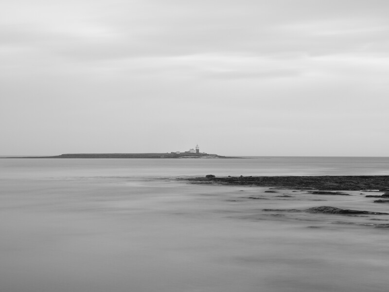 Minimalist seascape with distant lighthouse on rocky island under overcast sky.