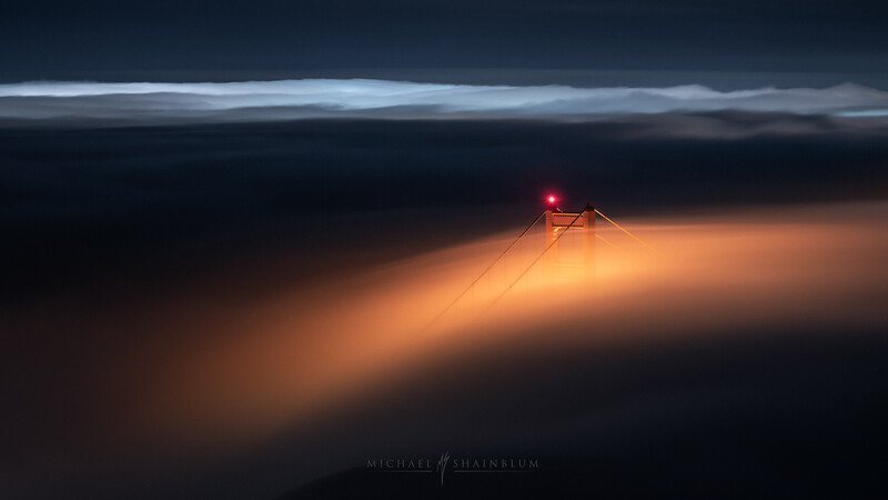 Illuminated radio or communication tower piercing through dense fog at dusk, with warm golden light rays spreading across the misty landscape.