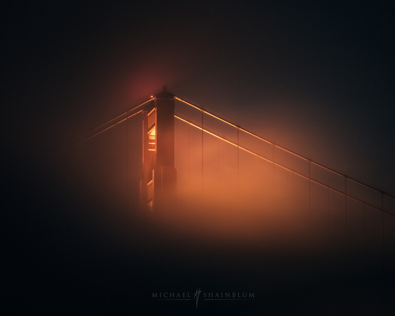Golden Gate Bridge tower emerging from thick fog at night, illuminated in warm orange light against a dark sky.