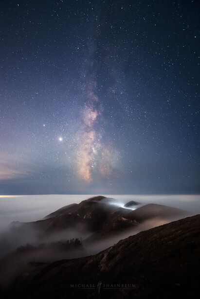 Milky Way galaxy arching above a mountain shrouded in fog beneath a starry night sky.