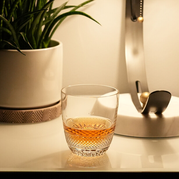 Glass of whiskey on a minimalist desk with potted plant and desk accessories in soft, warm light.